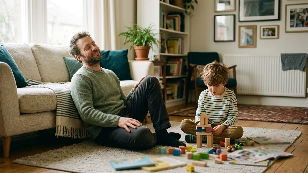 A tired father rests against the sofa while his young child plays with wooden blocks nearby